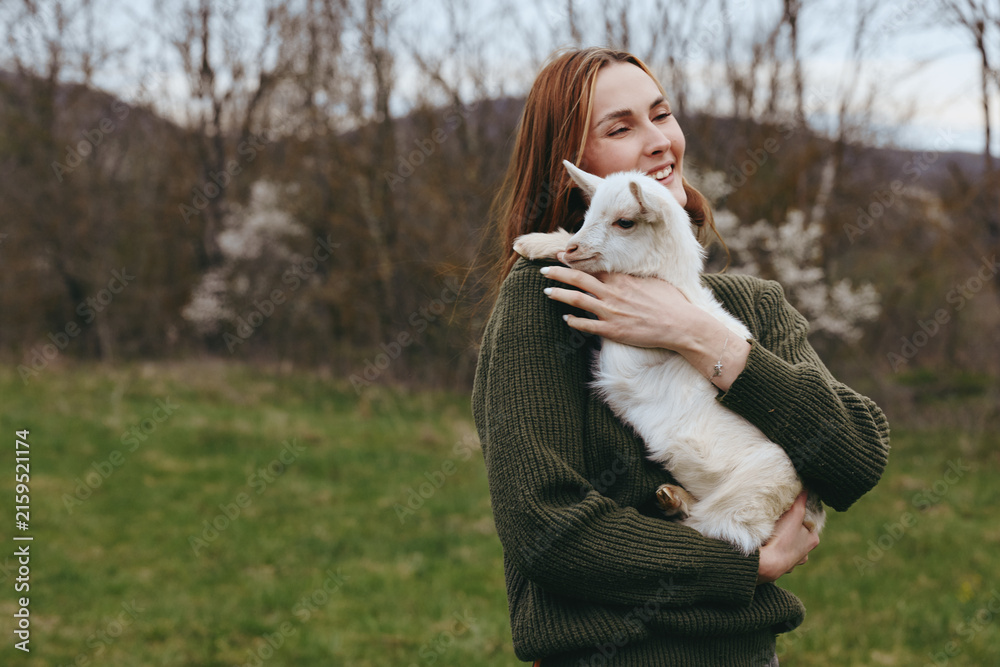 Naklejka premium A joyful woman in a knitted sweater hugs a white goat kid in a grassy countryside, capturing a simple lifestyle moment outdoors, with soft light and fresh air. The scene conveys affection and bond