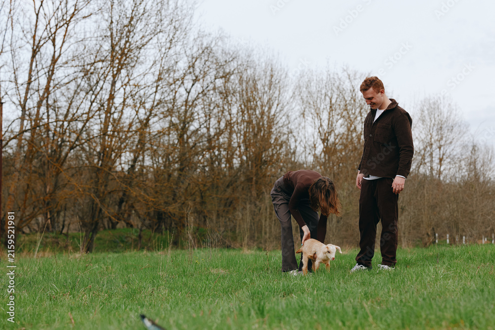 Naklejka premium Casual lifestyle scene with two adults in casual clothes in a grassy field, bending to greet a small light dog, during an autumn afternoon in a tranquil park setting, with soft natural light leaves