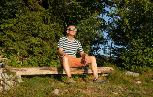 An older man wearing solar eclipse glasses is sitting on a wooden bench in the woods and watching a solar eclipse