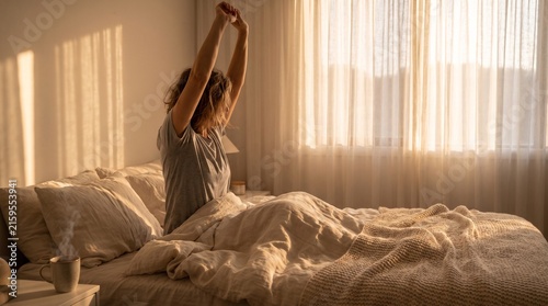 Woman waking up in bed stretching her arms in morning sunlight with sunlight
