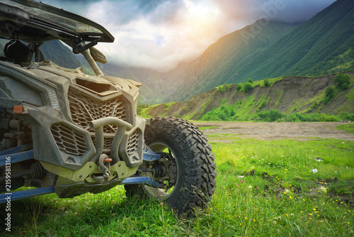 Сlose up of a yellow dirty offroad UTV buggy car parked on a dirt trail looking towards a majestic green mountain valley under a cloudy sky. Extreme motorsport travel adventure concept design