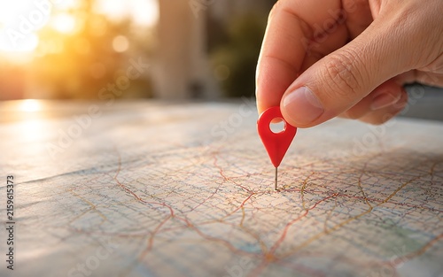 Hand holding red location pin marker on old paper map showing detailed street network and pointing to destination with warm sunlight.