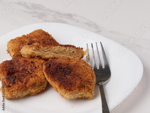 Fried, golden-brown soy cutlets served on a white plate with a fork. One of the cutlets is sliced ​​open, revealing the fibrous texture of the vegan meat substitute. A bright, high-key food photograph