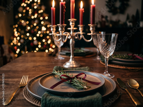 set christmas dining table with an elegant silver candelabra with burning red candles, a plate decorated with rosemary, and a decorated tree in background