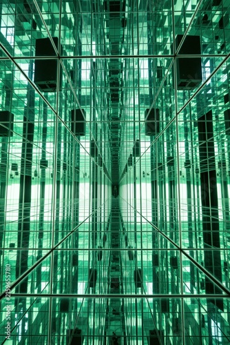 Infinite mirror room creates green light reflections. Geometric glass panels form futuristic tunnel effect. Abstract interior design space provides optical illusion. Disorienting environment offers