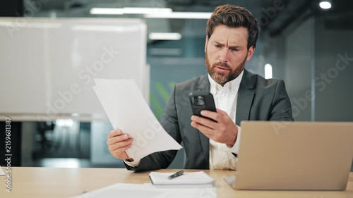 Professional experiencing confusion while comparing documents and smartphone data in modern office environment. Deep in thought over business information, seeking solutions problem solving.