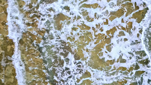 Fotografía Aerial view of waves crashing against a rocky coastline, creating a dynamic and foamy scene