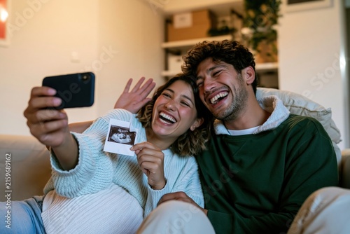 Happy couple sitting on a sofa making a video call and holding an ultrasound print, showing elated expressions for pregnancy announcement and family app marketing