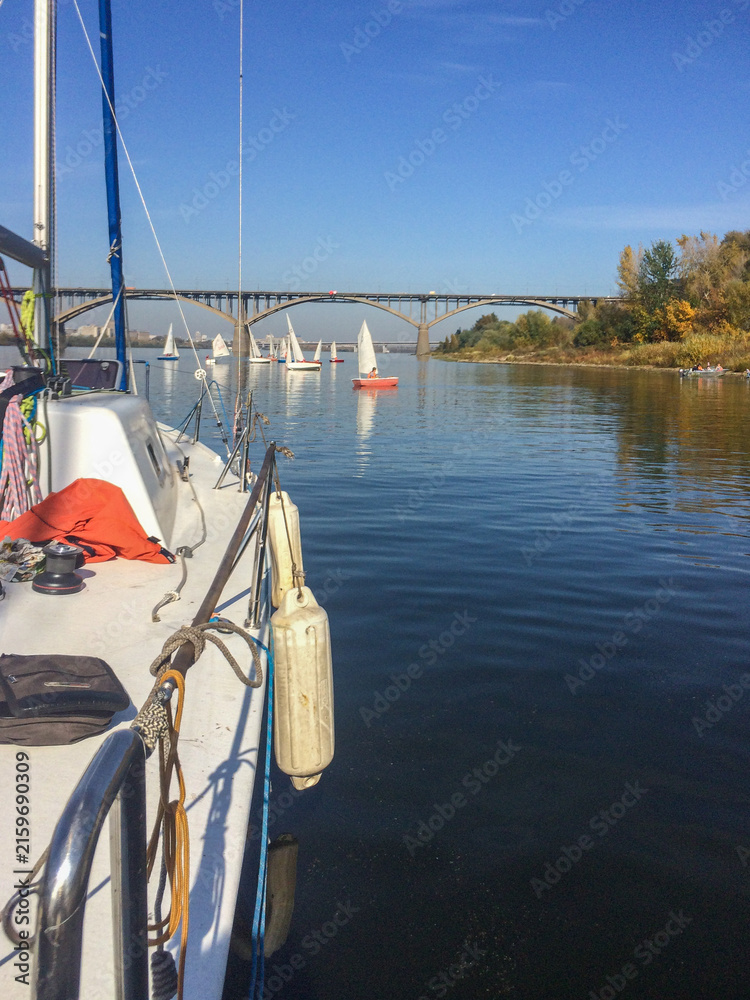 Fototapeta premium Sailing boats on a calm river near an arched bridge, view from the deck of a yacht.