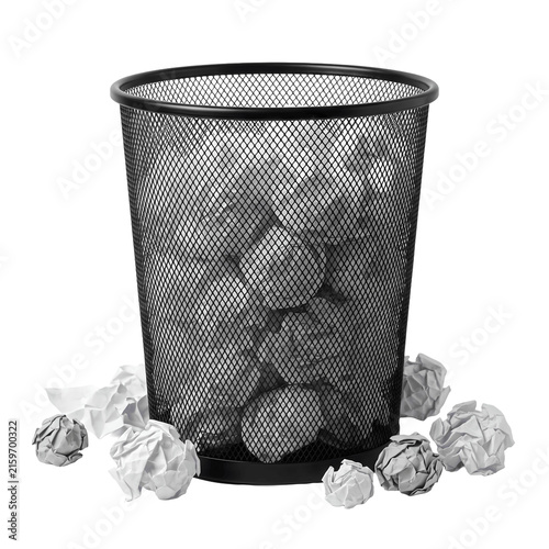 Black mesh wastebasket full of crumpled paper, with more discarded paper around it, isolated on white background