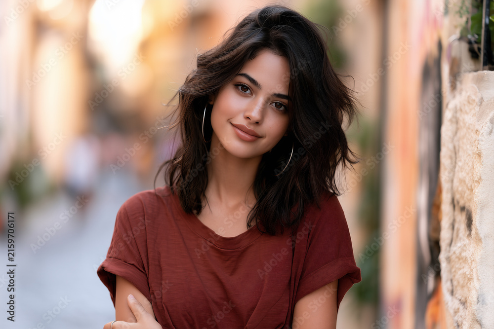 Obraz premium young woman with curly brown hair in a city alley, stylish brunette woman wearing casual clothes and silver hoop earrings, attractive young brunette standing outdoors in an urban setting