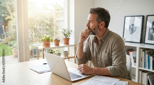 Middle aged man sitting at home office desk working on laptop computer while looking thoughtful at window view during remote work and career planning