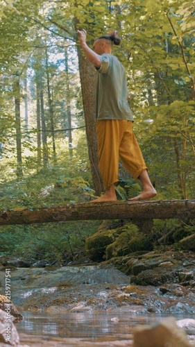 A man performs a balancing pose on a log in a serene forest setting. Dressed in light clothing, he focuses on maintaining his balance amidst lush greenery, embodying a zen-like state.