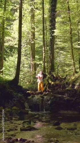 A person practices yoga on a moss-covered rock by a flowing stream in a serene forest, surrounded by lush greenery and dappled sunlight, creating a tranquil atmosphise.