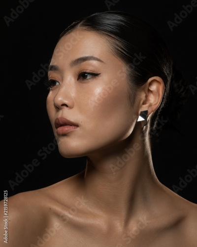 Stunning portrait of an Asian woman with sleek wet hair, dewy glowing skin, sharp winged eyeliner, and modern geometric silver earrings against a deep dark background