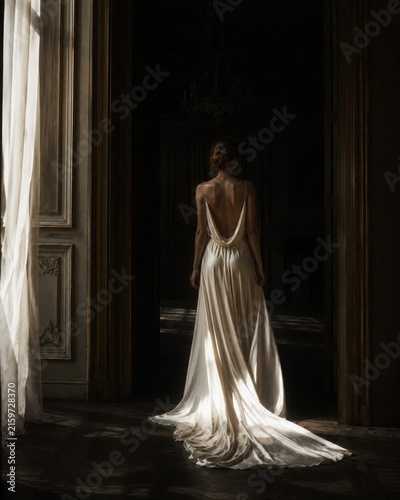 Sensual backless ivory silk slip dress on a graceful woman standing in a dark historic mansion illuminated by soft morning window light with long flowing train