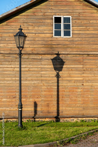 Obraz na plátně Vintage street lamp casting a long distinct shadow across a sunlit weathered wooden wall in the early morning