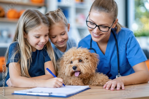 Female Caucasian veterinary nurse completing pet forms with two girls and a curly dog, ideal for clinic registration, family pet care and animal health education.