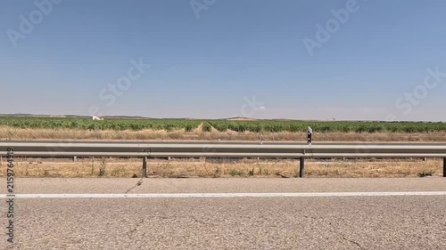 A lateral view of a highway with a metal guardrail, surrounded by fields of green crops and dry grass under a clear blue sky. The video captures the essence of rural road travel in Spain
