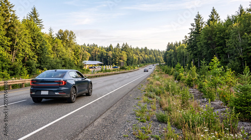 A modern dark sedan driving on a scenic highway surrounded by lush green pine forests.