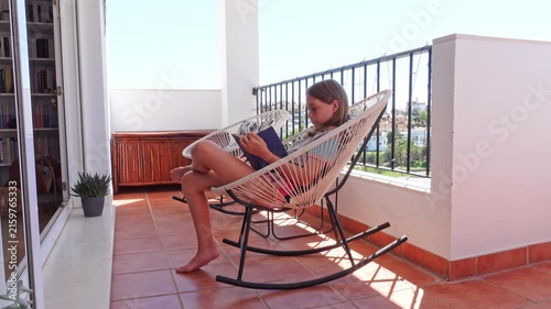 Preteen girl reading a book sitting and relaxing in a modern rocking chair on a terrace on a sunny summer day
