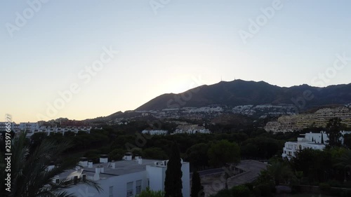 Time-lapse of a serene sunset over a horizon mountainous landscape with residential buildings and lush greenery, with clear sky. Sun moving fast
