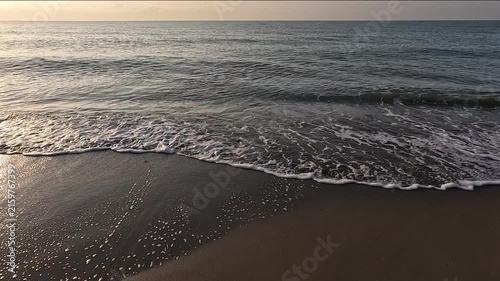 Waves of a calm Mediterranean sea reach the brown sand beach, with a horizon line at sunrise or sunset with a clear sky
