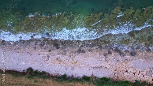 The camera captures an aerial view of a rugged coastline, where waves crash against rocky shores. A lone figure, gender indistinct, walks along the beach, immersed in nature's serene beauty.