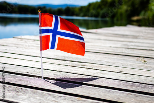 Small Norway flag beside calm water on rustic pier under clear sky evoking Scandinavian identity patriotism tourism and easygoing summer leisure with soft bokeh and space for text