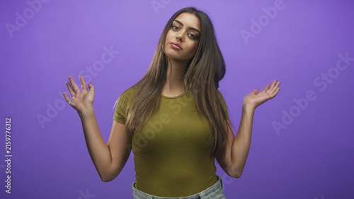 Obraz na plátne Woman of hispanic appearance with hands raised palms up in a shrug gesture wearing olive shirt and jeans against purple studio backdrop; indifference casual