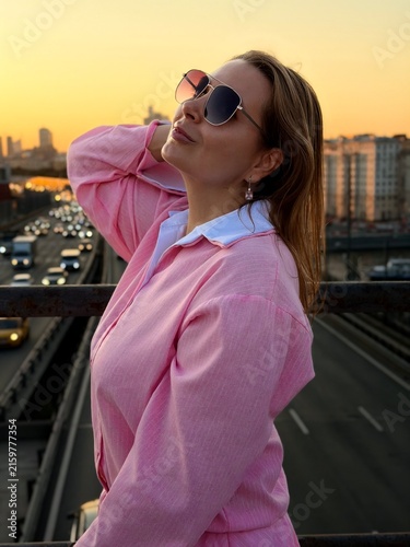 Woman in sunglasses and pink shirt stands on a bridge at sunset, with a city skyline and busy road filled with cars in the background