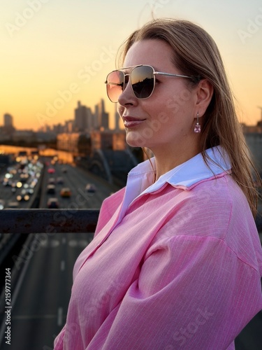 Woman in sunglasses and pink shirt stands on a bridge at sunset, with a city skyline and busy road filled with cars in the background