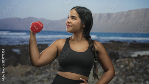 Cuadro en lienzo Young woman holding red 3kg dumbbell with bent arm on rocky beach, wearing black sports bra and braid; confidence strength motivation