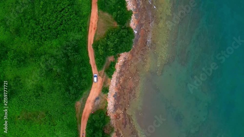 A lone traveler, seen from an aerial view, drives along a winding dirt road beside a rocky coastline. The lush green forest contrasts with the turquoise waves crashing against the shore.