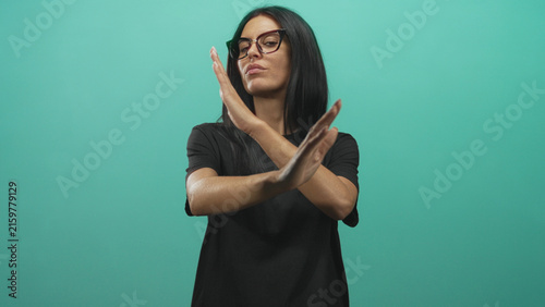 Bild auf Leinwand Woman hands crossed in x stop gesture, wearing glasses and black t shirt against turquoise studio backdrop; defiance