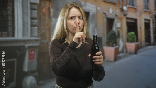 Foto Woman holding beer bottle, finger to lips for silence on a narrow cobblestone street with planters and windows; warning secrecy caution