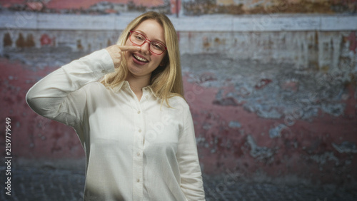 Woman touching nose with finger in a street setting by a weathered wall, wearing red glasses and white shirt; playful mischief.