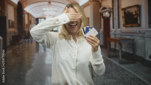 Woman holding creditcards and smiling while sometimes covering eyes with her hand in an ornate building hallway; joy financial confidence.