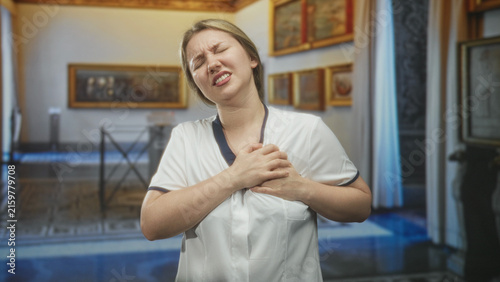 Fotografie Woman in white uniform clutching chest in museum, grimacing and pressing hands over sternum suggesting heartattack and urgent pain; medical emergency distress