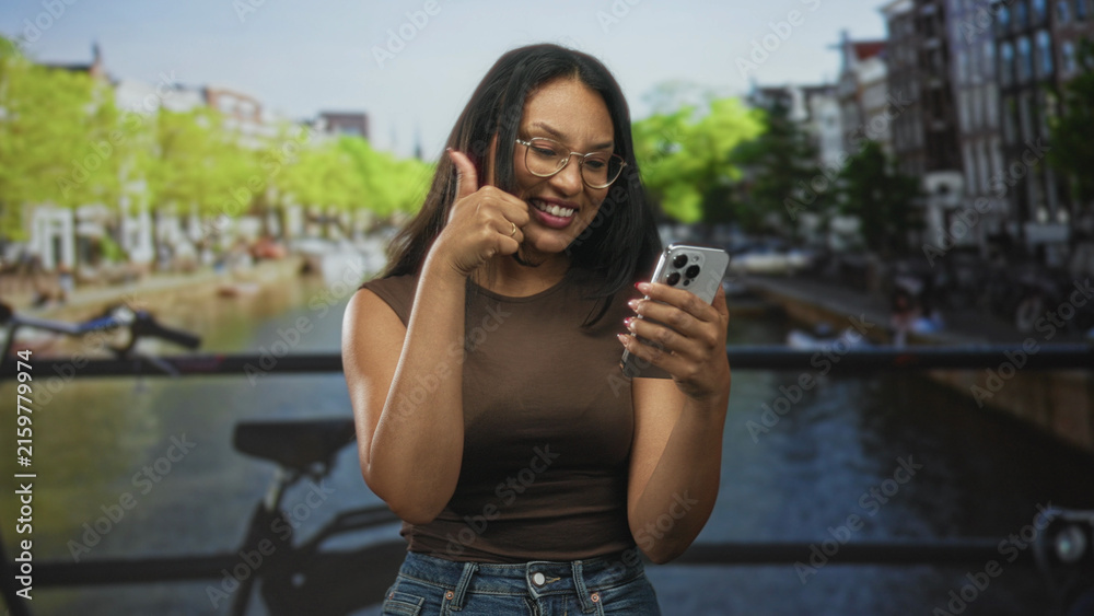 Fototapeta premium Woman tapping smartphone and giving thumbs up with hands visible on street canal bridge wearing glasses and jeans; digital connection happiness.