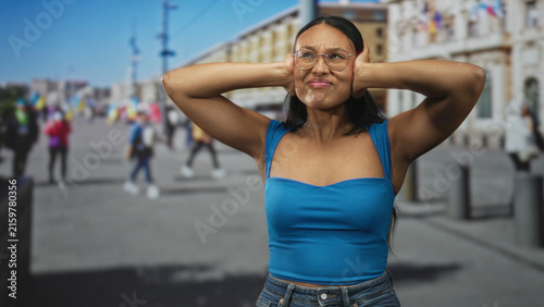 Bild auf Leinwand Woman in blue top tightly covers ears with both hands, eyes closed and grimacing on a busy urban pedestrian street outside a large neoclassical building; sensory overload distress