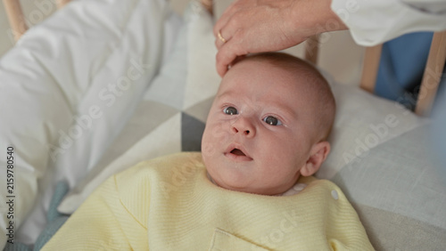 Quadro su tela Newborn baby in yellow onesie lies in crib on patterned pillow as a hand gently pats forehead beside a hanging mobile in nursery; calm bonding