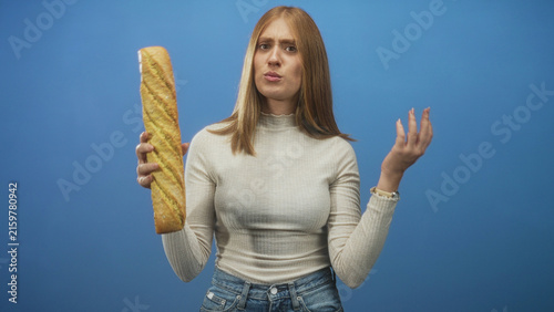 Фотография Young redhead woman holds baguette in one hand and gestures with open palm against blue studio wall; confusion