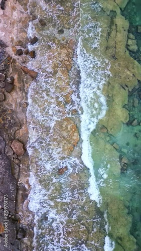 A serene coastline unfolds in this aerial view, where gentle waves caress the rocky shore. The clear waters reveal hidden rocks beneath, offering a peaceful glimpse into the seaside's natural beauty.