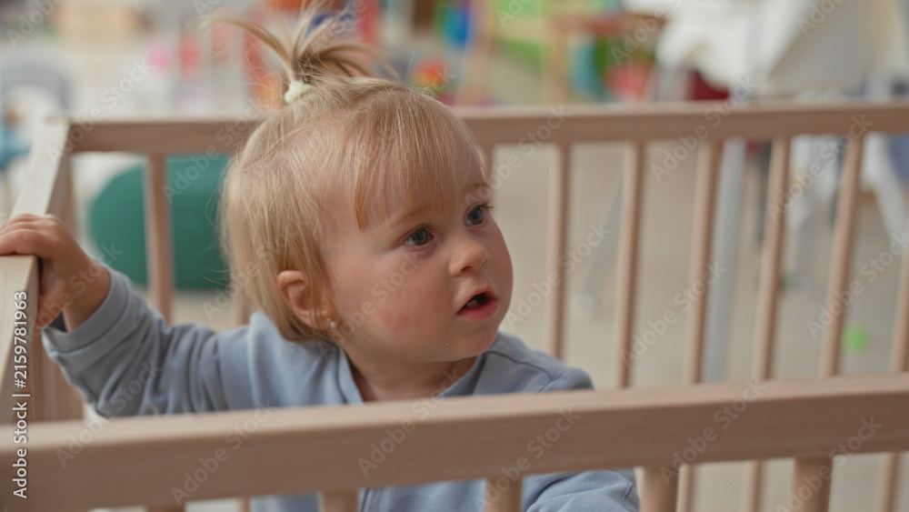 Fototapeta premium Toddler girl with blonde ponytail grips wooden crib rail with small hands, looking sideways near play area in building; quiet curiosity.