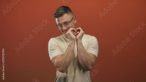 Quadro su tela Young man with glasses and short blond hair forms heart with hands over chest while smiling in studio; playful warm affection
