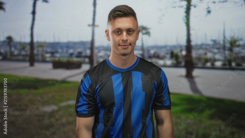 Naklejka premium Man smiling with visible face and tilted head wearing blue soccer jersey on street promenade by marina boats, hands relaxed at sides; pride team spirit.