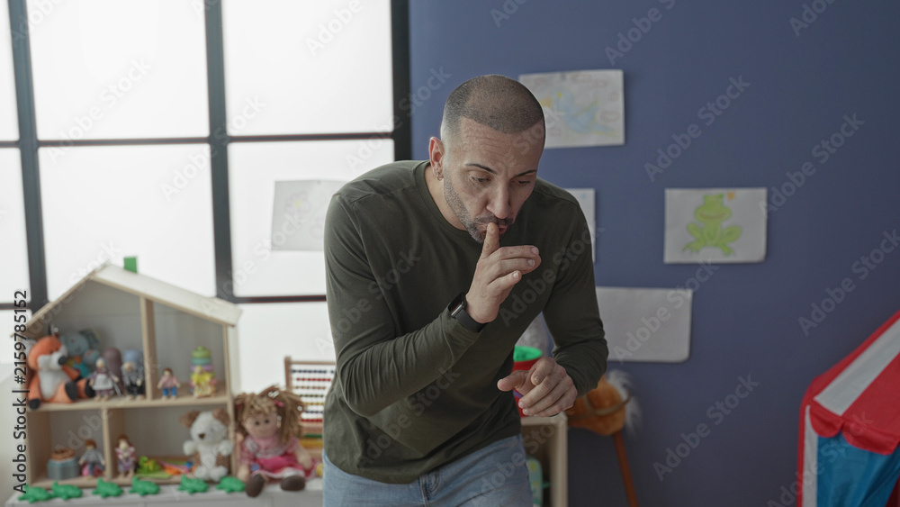 Fototapeta premium Man with hands gesturing and finger to lips for silence in a kindergarten playroom in a school building; patience classroom guidance.