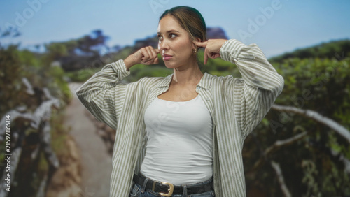 Konstfotografi Woman plugs ears with index fingers on forest path, wearing white tank top and striped overshirt, eyes closed and lips pursed; annoyance
