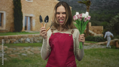 Canvas Print Woman gardener in red apron holds hand trowels and a bouquet of tulips while grimacing in front of building; frustration resilience